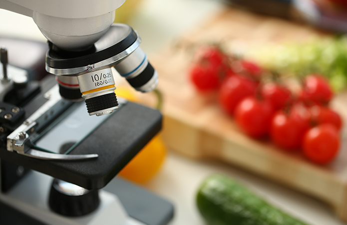 Microscopio in laboratorio con verdure fresche sullo sfondo, rappresentativo di analisi alimentare e sicurezza microbiologica, simbolo dell'articolo del blog food di Lifeanalytics sui nitriti e nitrati negli alimenti: sicurezza, limiti e novità normative