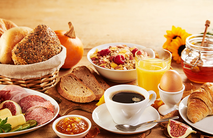“Ricca colazione con caffè, succo d’arancia, muesli, pane, salumi, formaggi, uova e croissant su tavolo rustico, simbolo dell'articolo del blog che tratta la tematica delle Direttive colazione e perchè la loro revisione è fondamentale per produttori e consumatori, relativo alla business unit food di Lifeanalytics 