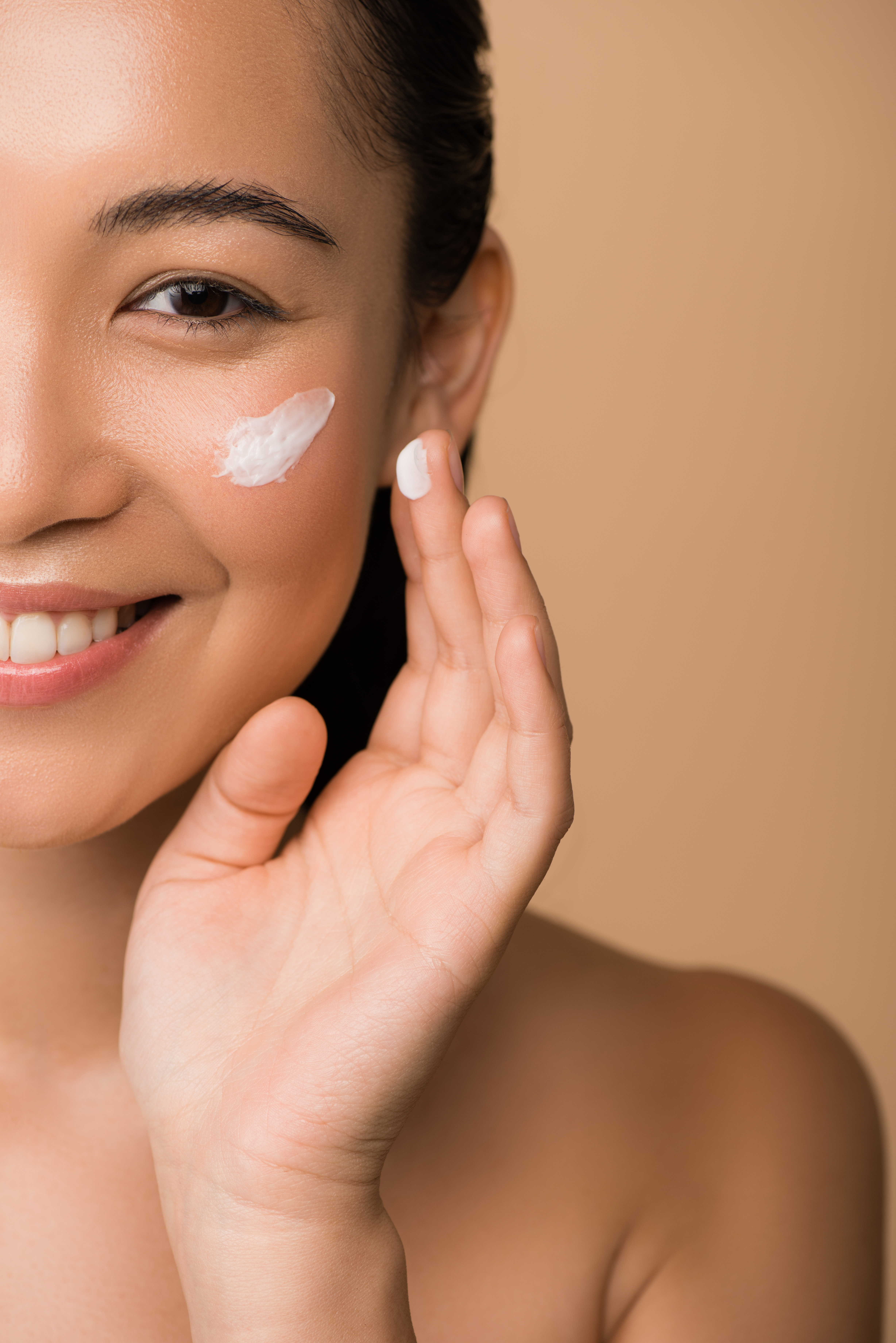 Donna sorridente che applica crema idratante sul viso su sfondo color beige simbolo dell'articolo del blog che tratta la tematica della luce blu emessa dagli schermi di smartphone, computer e lampadine LED e del suo potenziale impatto sulla pelle e sulla salute generale, relativo alla business unit cosmetic & medical device di Lifeanalytics