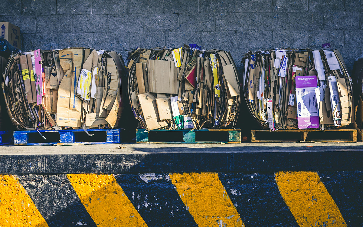 Immagine mostra balle di cartone compresse su pallets, ordinate contro un muro di cemento e sopra un marciapiede decorato da strisce gialle e nere, simbolo del regolamento su trasporto si merci su strada