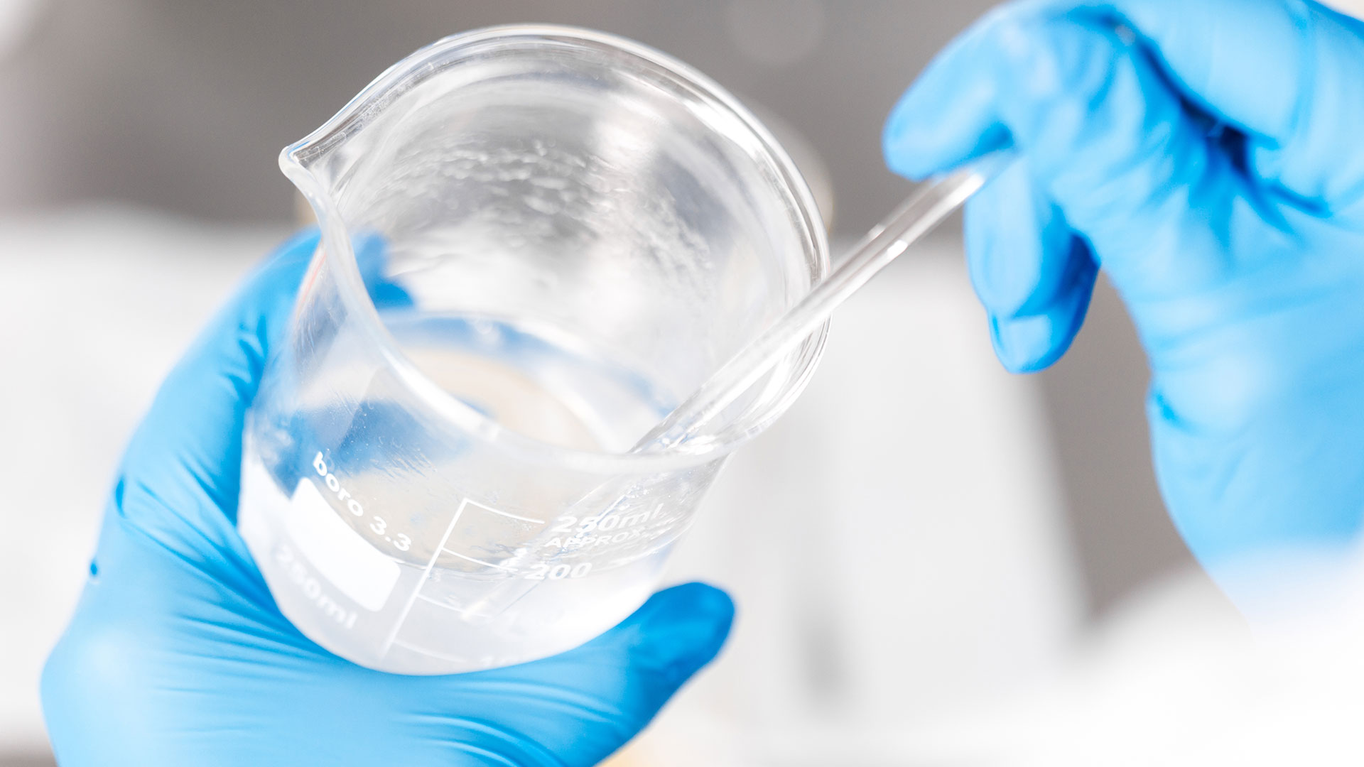 Mani con guanti blu che maneggiano un becher di vetro trasparente con liquido incolore, utilizzando una pipetta per analisi chimiche si laboratorio, su sfondo neutro e luminoso