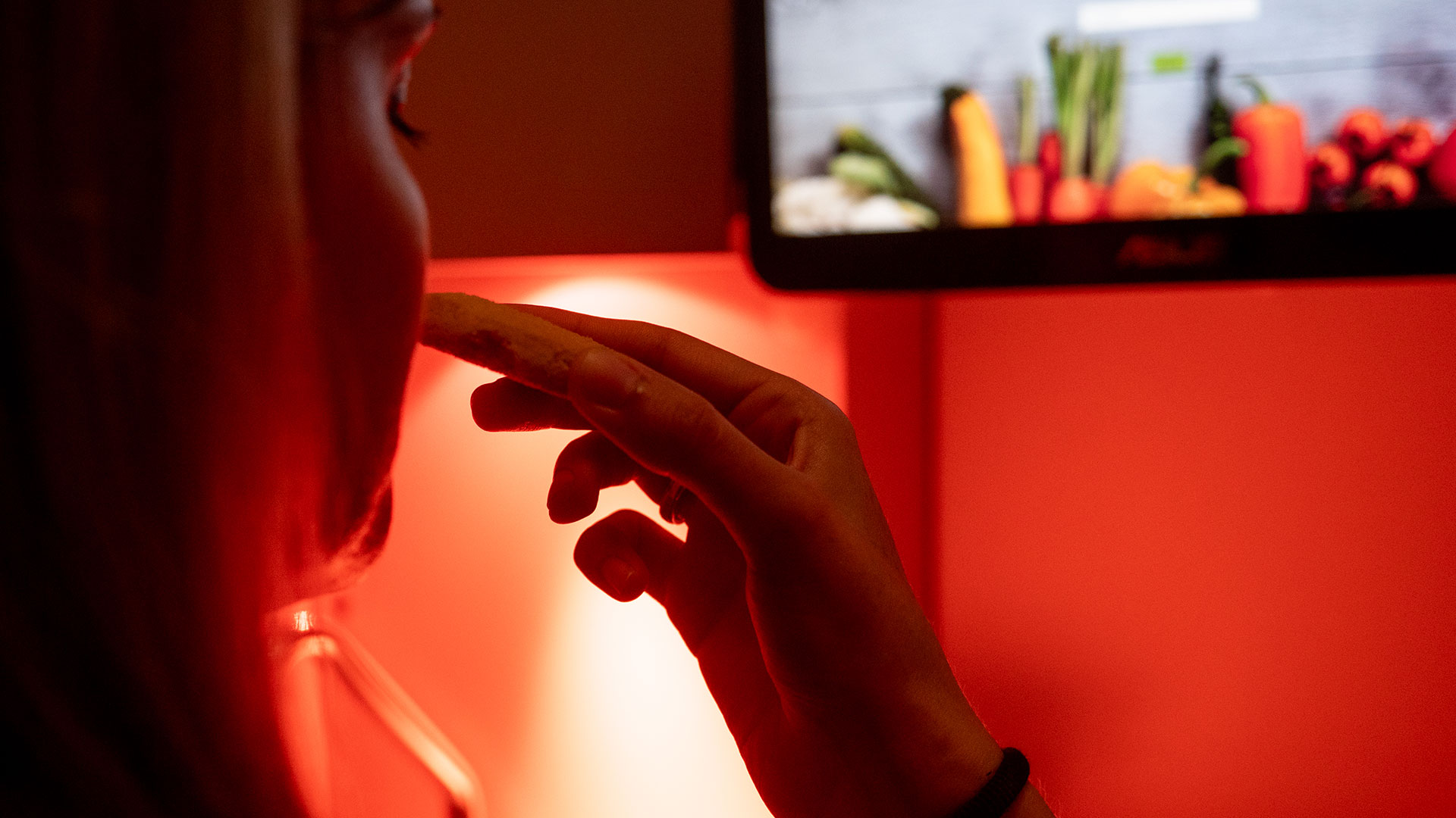 Persona che assaggia del cibo in un ambiente illuminato di rosso con uno schermo che mostra verdure sullo sfondo, simbolo delle analisi sensoriali 
