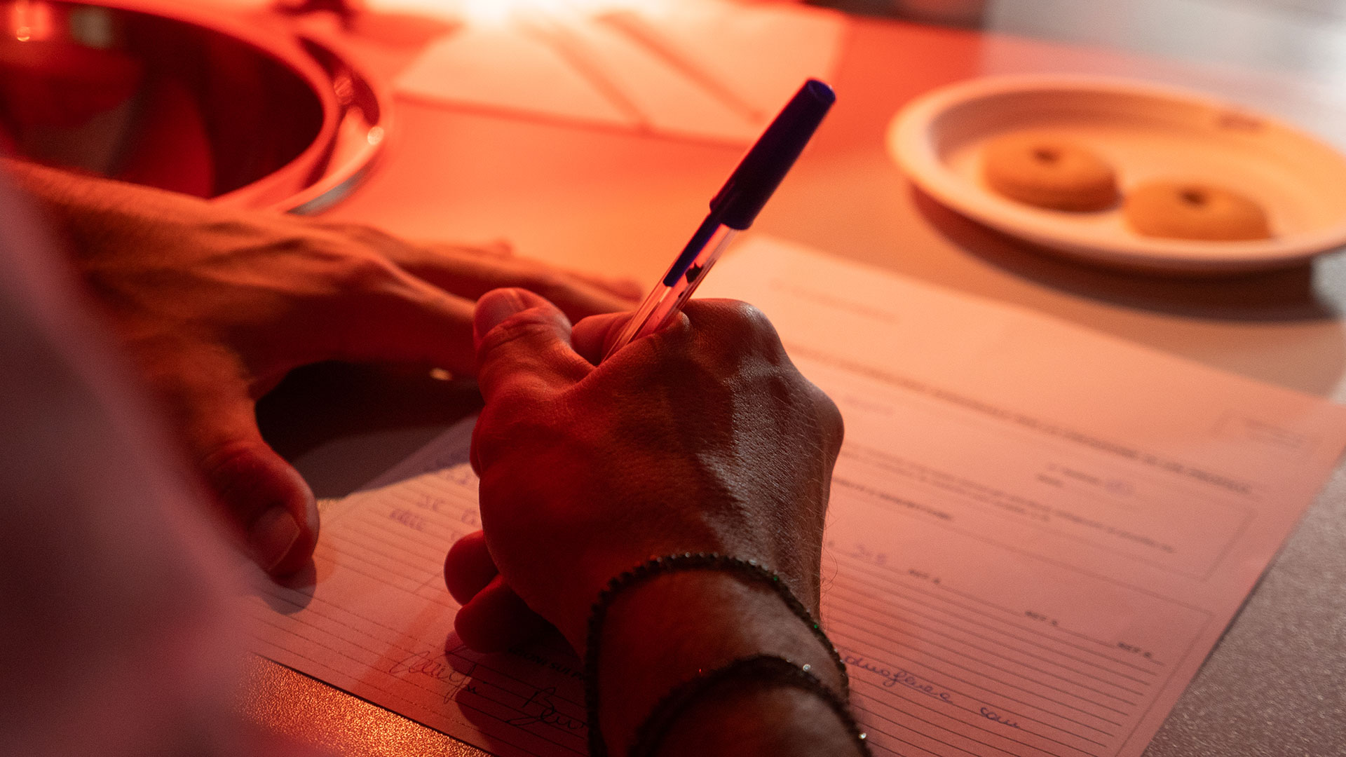 Mano che scrive su un modulo con una penna in un ambiente illuminato di rosso, sul tavolo un piatto con biscotti, simbolo delle metodologie utilizzate per le ricerche qualitative ed esplorative