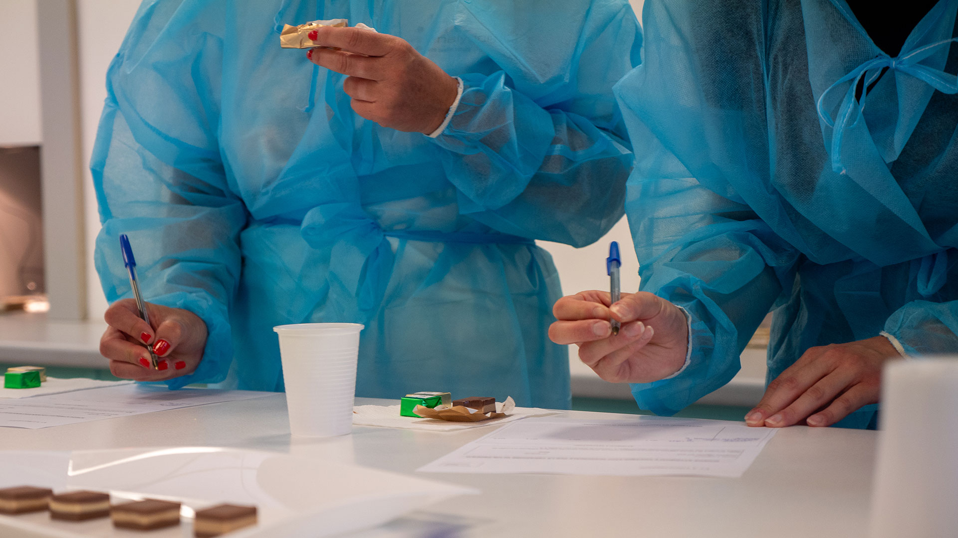 Persone in camici blu monouso che scrivono su moduli e assaggiano campioni di cibo durante un'analisi sensoriale, simbolo dei metodi descrittivi e discriminanti per conoscere un prodotto