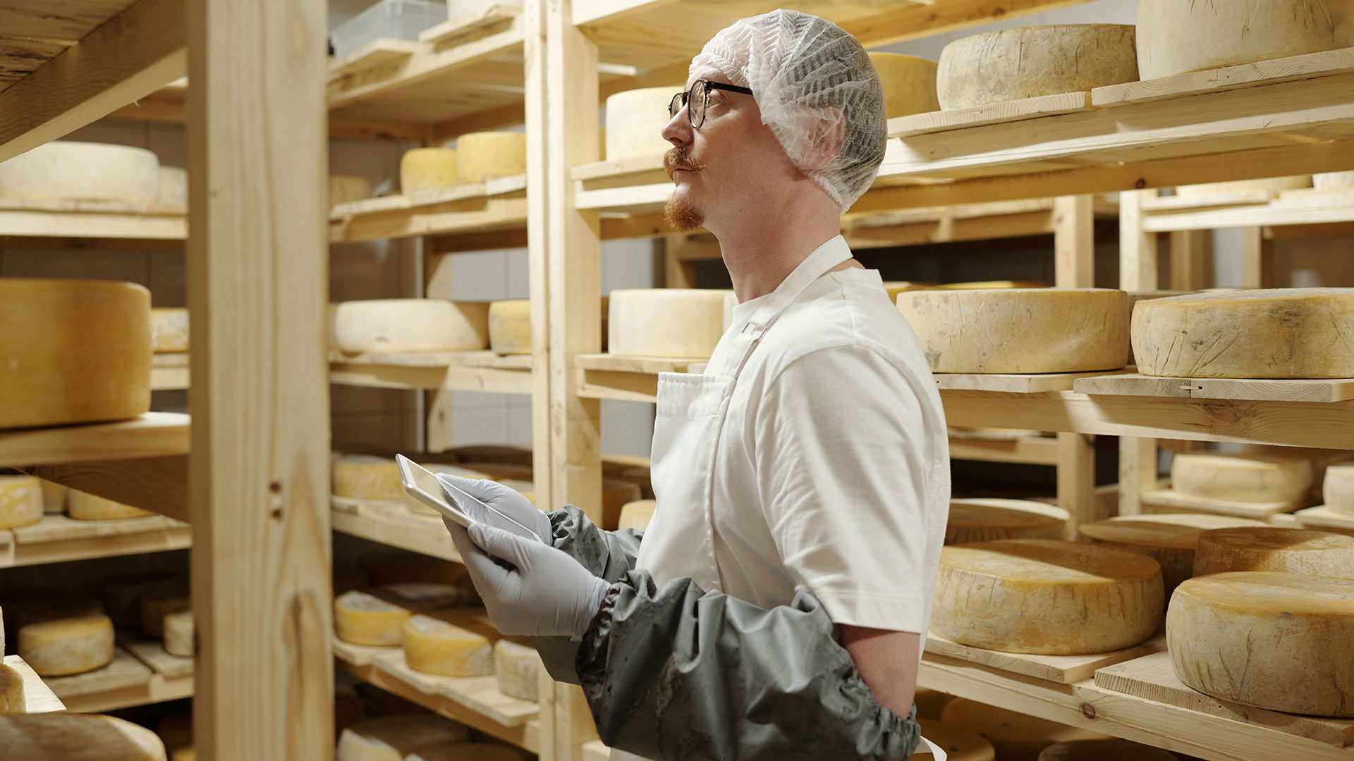 Immagine di tecnico alimentare con grembiule e protezioni che sta visionando delle forme di formaggio con un tablet per prendere appunti, simbolo del servizio dedicato agli audit interni e di seconda parte