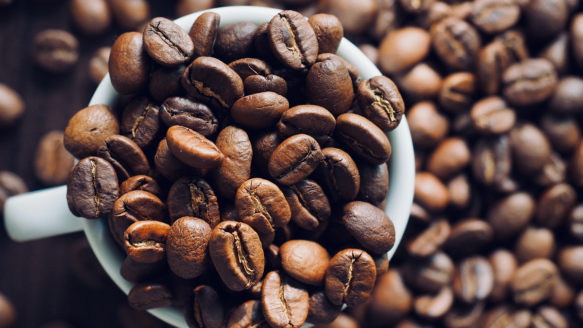 Tazza bianca piena di chicchi di caffè tostati, con altri chicchi sparsi sullo sfondo, simbolo del servizio Caffè, olio, uova, cacao e grassi alimentari della business unit food di Lifeanalytics