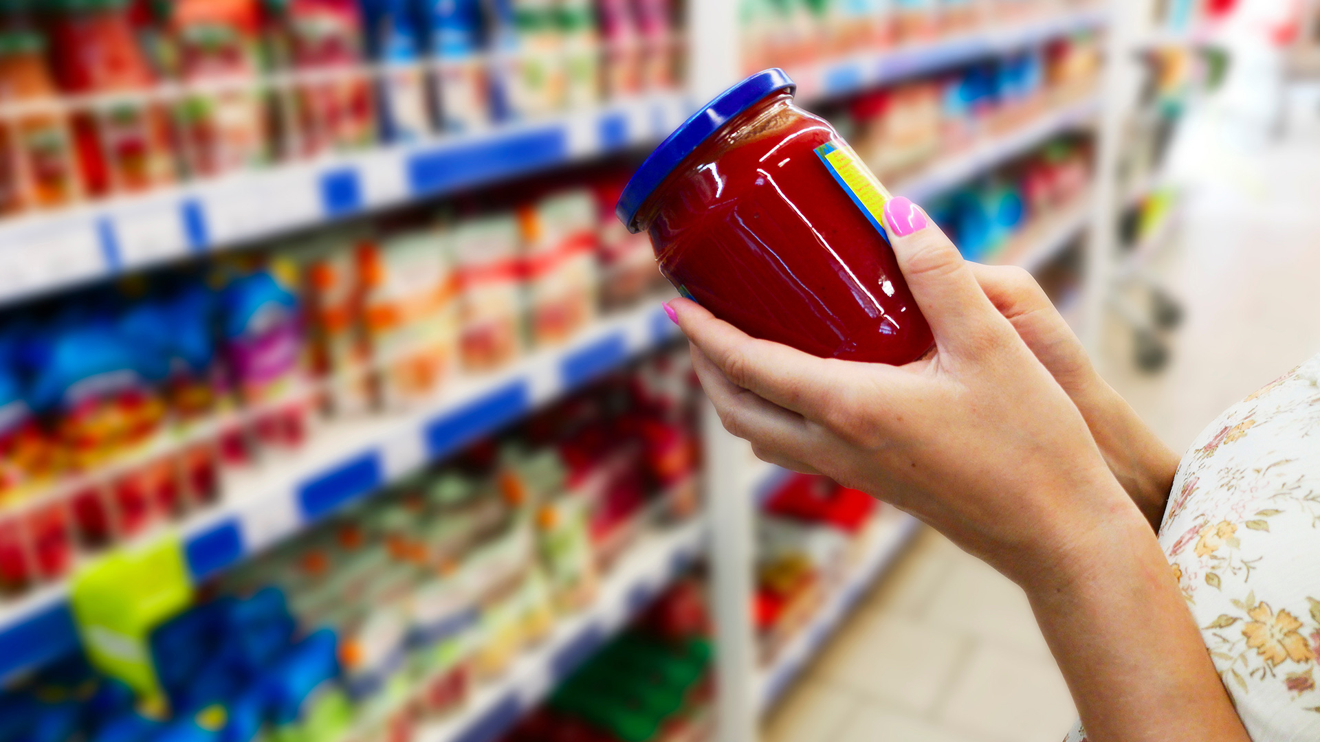 Donna che tiene in mano un barattolo di salsa di pomodoro in un supermercato, osservando l'etichetta davanti allo scaffale dei prodotti alimentari, simbolo del servizio food ortofrutta e conserve di Lifeanalytics