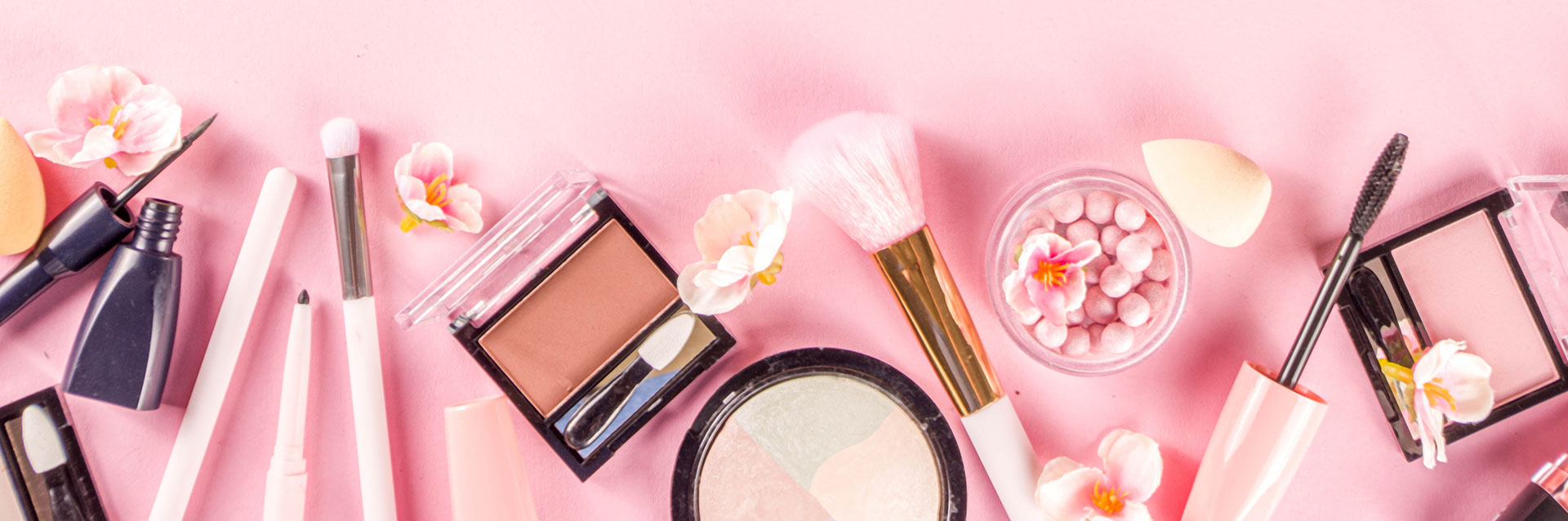 Collezione di prodotti di makeup disposti su uno sfondo rosa, inclusi mascara, pennelli, eyeliner, blush e rossetti, decorati con delicati fiori rosa, che incarnano un'estetica fresca e femminile