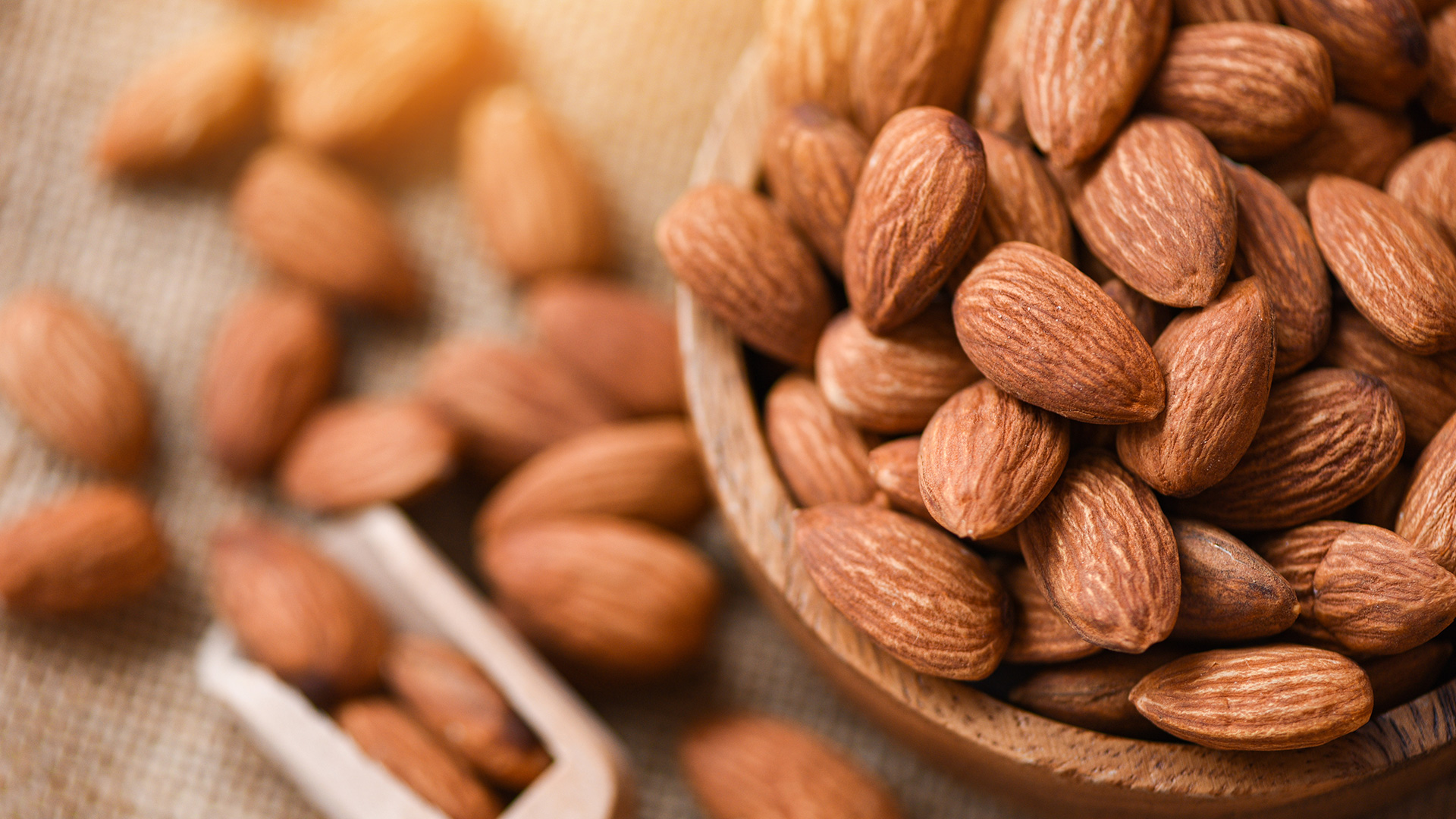 Shelled almonds in a wooden bowl and scattered on a jute surface, symbolizing Lifeanalytics' dried fruit and tree nuts service