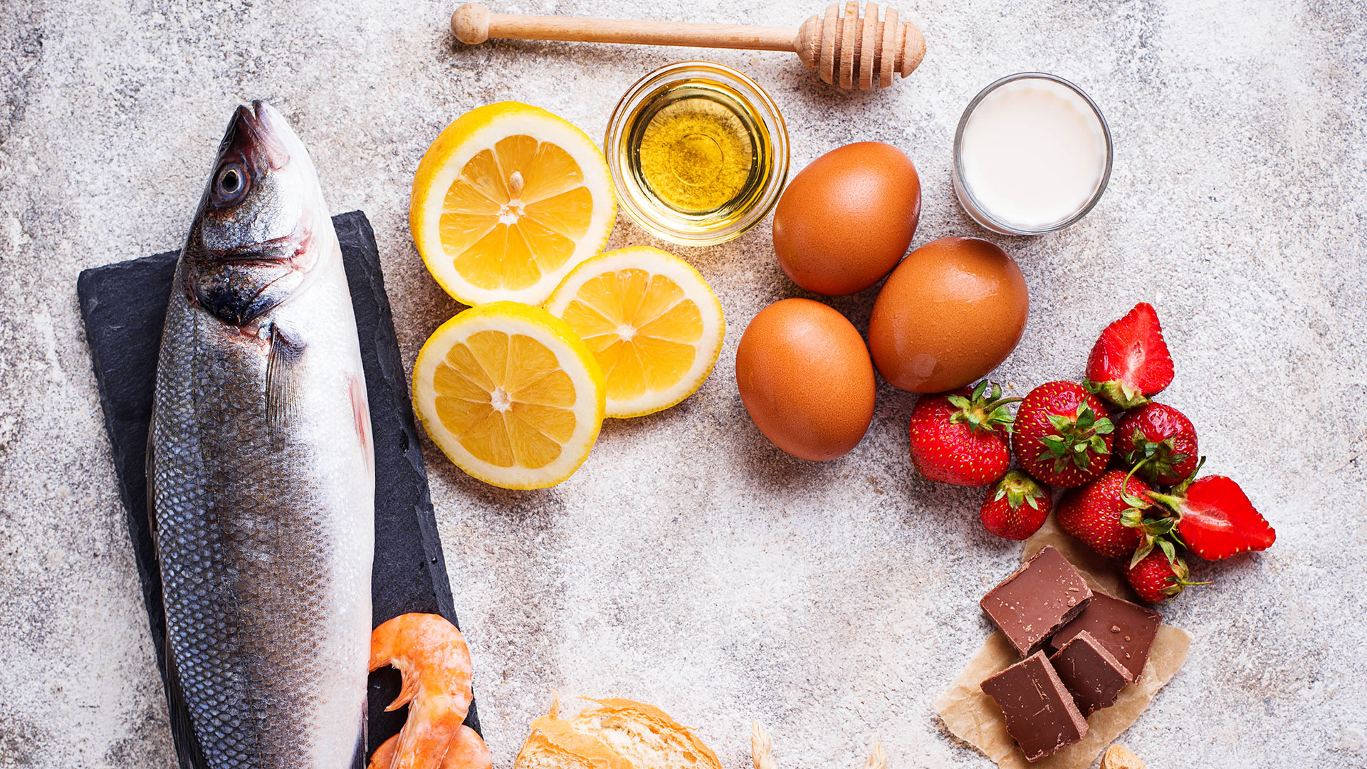 Immagine di un piano da lavoro con sopra alimenti quali pesce, limone, miele, fragole, uova, cioccolato, gamberetti e latte, simbolo del corso dedicato alla gestione del rischio allergeni