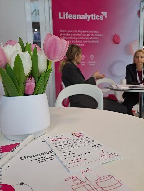 Primo piano del desk presente in fiera con brochure e flyer Cosmetic and Medical device con un vaso di tulipani bianchi e rosa e sullo sfondo le pareti dello stand con il colore caratteristico di questa business unit, simbolo dell’articolo del blog dedicato alla fiera Lifeanalytics a in-Cosmetics Global 2026: direzioni future nell’ambito della business unit cosmetic & medical device di Lifeanalytics
