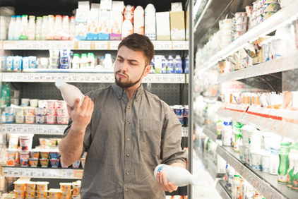 Uomo giovane al supermercato mentre confronta due bottiglie di latte davanti a uno scaffale refrigerato ricco di prodotti lattiero-caseari, simbolo dell'articolo del blog della business unit Food dedicato all'attenzione alle etichette con dicitura “artigianale” negli alimenti e cosa cambia con la nuova normativa