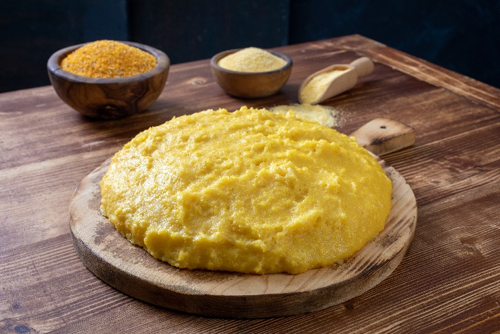 Polenta tradizionale di mais gialla disposta su un tagliere rotondo in legno, con ciotole contenenti farina di mais e una paletta sullo sfondo, su tavolo rustico, simbolo dell'allerta segnalata per il superamento dei limiti di aflatossine, micotossine prodotte da muffe che si sviluppano sui cereali in specifiche condizioni di stoccaggio e campo
