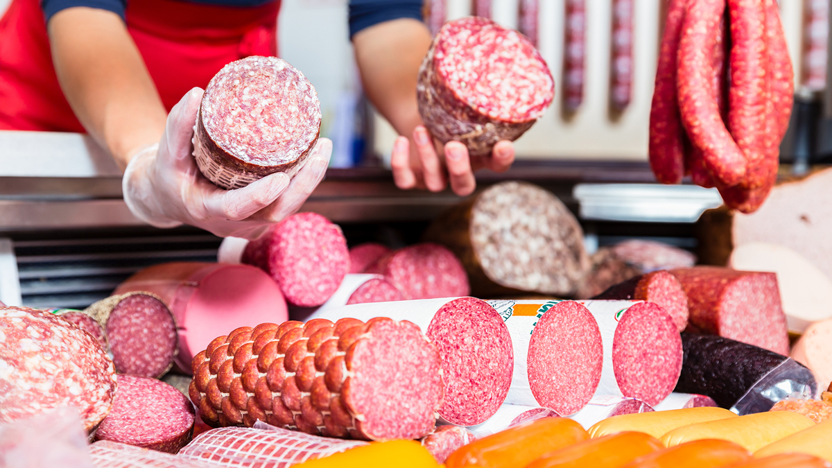 Banco di salumi assortiti con salami affettati e interi, esposti in una salumeria; operatore con guanti mostra due salami, simbolo del servizio carne e derivati della business unit food di Lifeanalytics