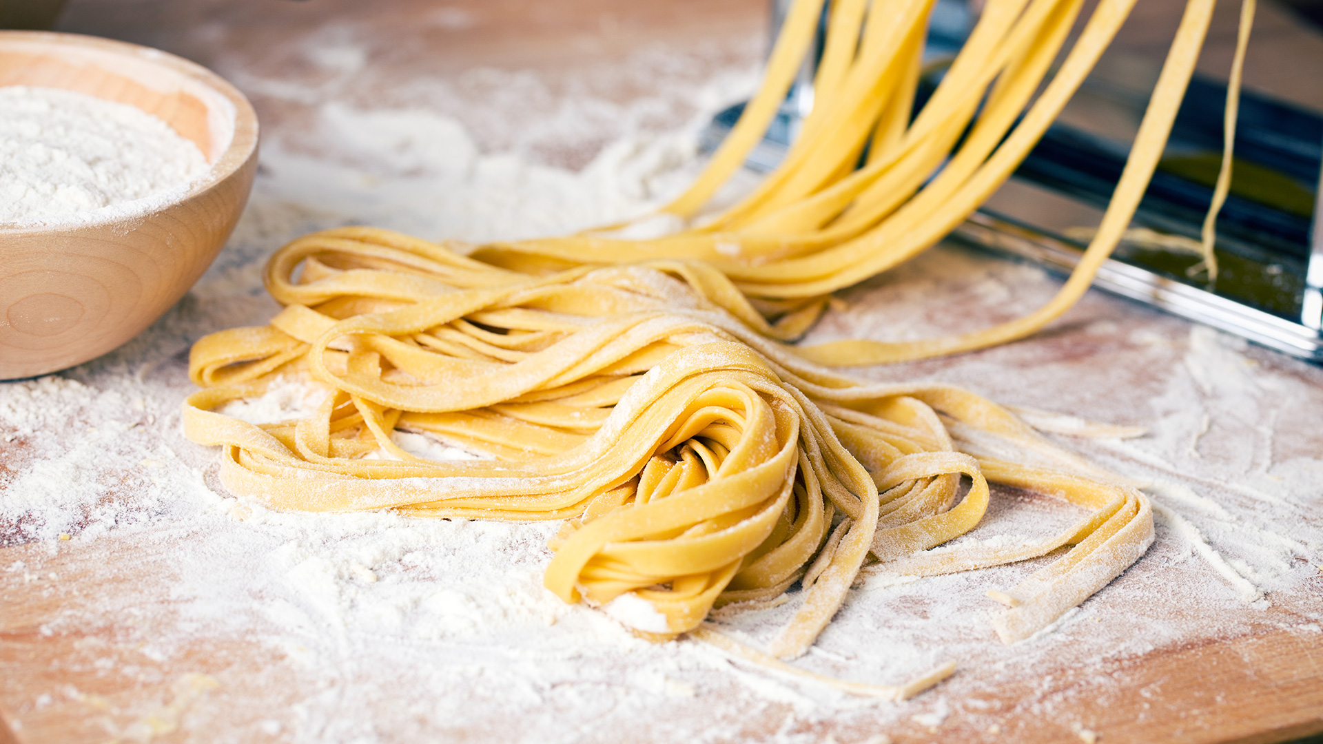 Fresh egg pasta cut by hand on a floured surface, with a bowl of flour and a pasta machine in the background, symbolizing Lifeanalytics' pasta and bakery products service