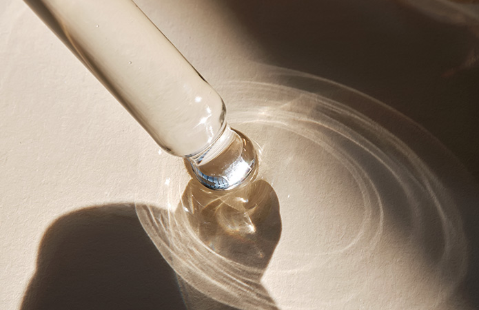rimo piano di una pipetta in vetro con goccia di siero trasparente su superficie chiara, illuminata da luce naturale, simbolo dei l’uso promettente dei peptidi in cosmetica e cosmeceutica
