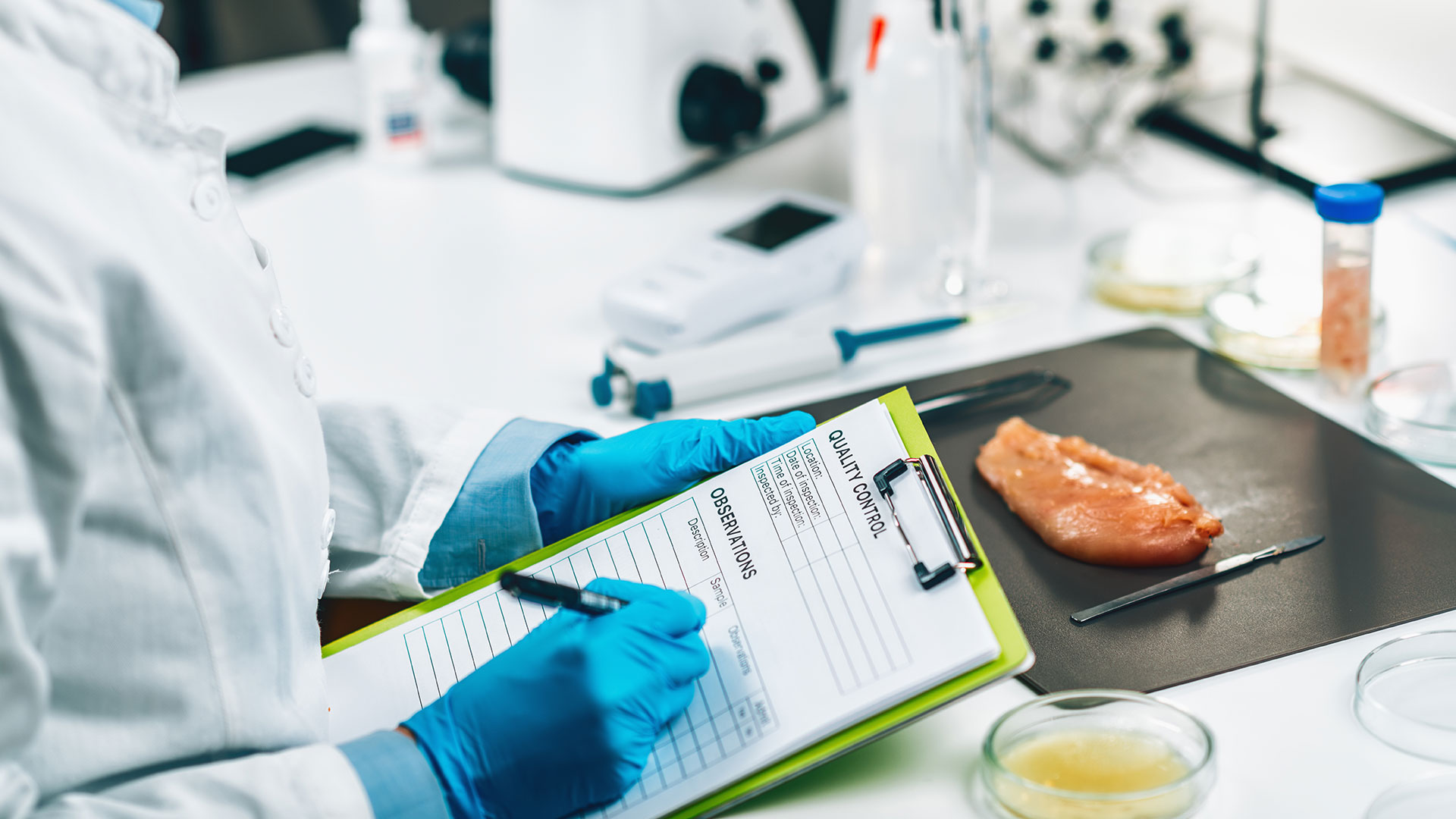 Techico di laboratorio con camice e guanti che sta prendendo appunti cimpilando una tabella di controllo qualità, davanti ad un tavolo di laboratorio con carne e strumenti in vista, simbolo del servizio dedicato alle perizie tecniche e relazioni specialistiche