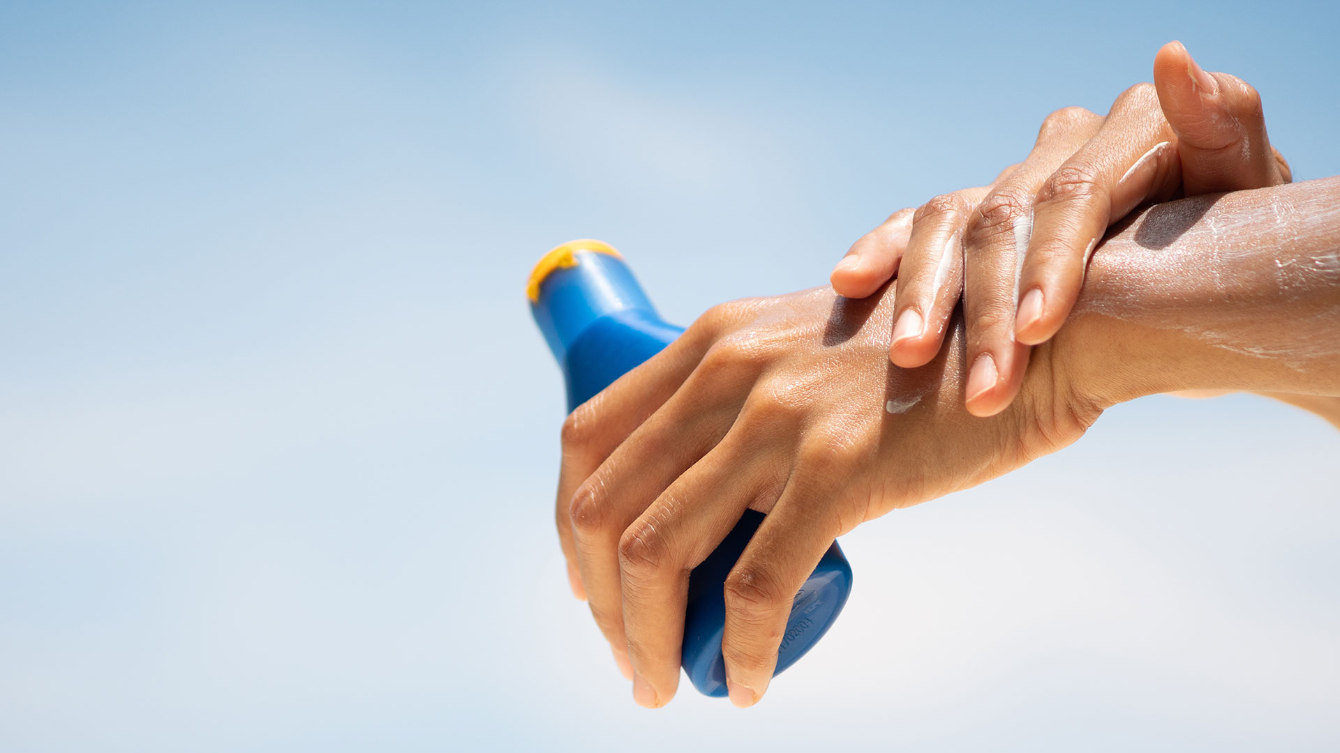 Applicazione di crema solare su una mano con flacone blu sotto il cielo estivo, simbolo del servizio Suncare della business unit cosmetic and medical device di Lifeanalytics