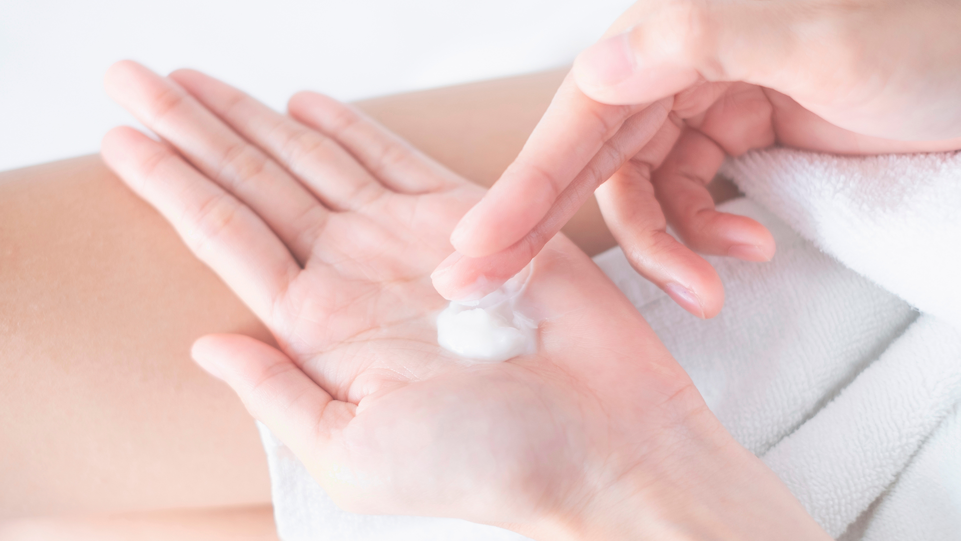 Mani che applicano crema idratante su palmo della mano, su sfondo chiaro con asciugamano bianco, simbolo del servizio skincare della business unit cosmetic and medical device di Lifeanalytics