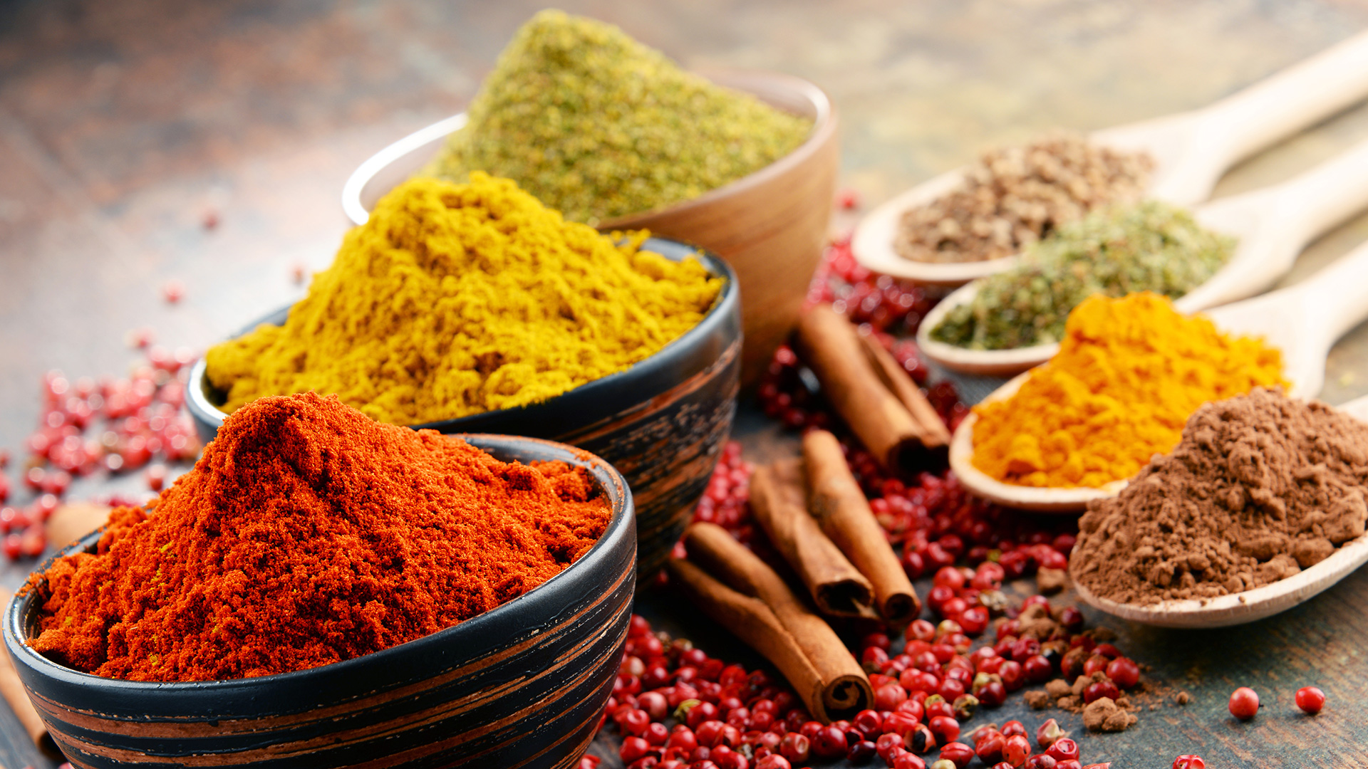 arietà di spezie in polvere di diversi colori in ciotole di legno e cucchiai, insieme a bacche di pepe rosa e stecche di cannella su sfondo rustico, simbolo del servizio spezie della business unit food di Lifeanalytics