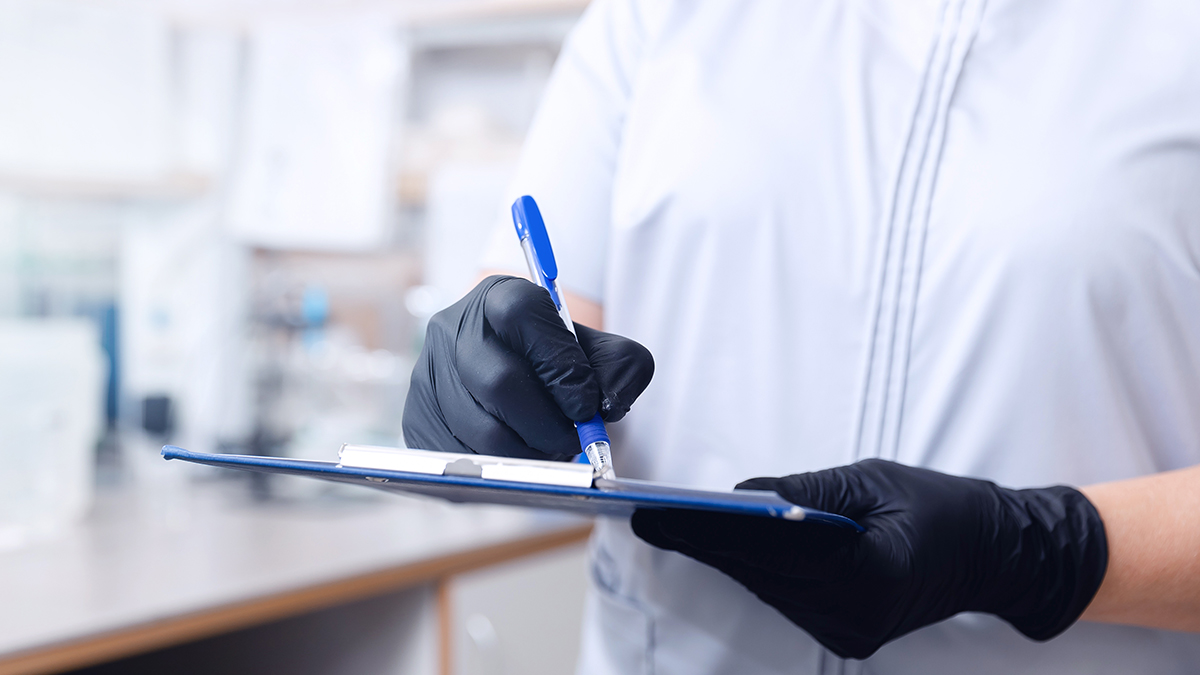 Operator in a white lab coat and black gloves writing on a clipboard with a blue pen inside a laboratory, representing the Haircare service of the cosmetic and medical device business unit of Lifeanalytics.