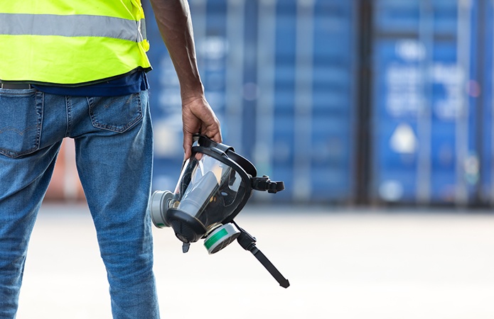 Operaio con giubbotto ad alta visibilità che tiene in mano una maschera antigas in un'area industriale, simbolo dell'articolo del blog che tratta la tematica dell'amianto e del radon, relativo alla business unit ehs di Lifeanalytics