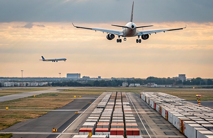 Aereo commerciale in fase di atterraggio sopra una pista aeroportuale con container di carico allineati, e un secondo aereo che si avvicina in lontananza al tramonto, immagine simbolo dell’articolo dedicato alle ultime novità sull'ADR 2025 e il Regolamento IATA per la business unit EHS di Lifeanalytics