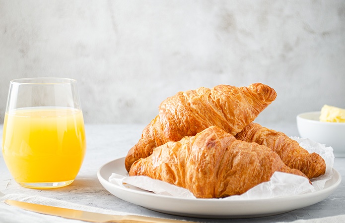 Colazione con croissant freschi su un piatto e bicchiere di succo d’arancia, simbolo dell'articolo del blog che tratta la tematica delle Direttive colazione e perchè la loro revisione è fondamentale per produttori e consumatori, relativo alla business unit food di Lifeanalytics 