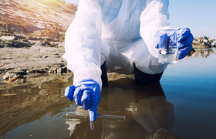 Tecnico ambientale in tuta protettiva e guanti che preleva campioni d’acqua da un fiume o lago per analisi di contaminanti, immagine simbolo dell’articolo dedicato a come valutare e gestire i siti potenzialmente contaminati  per la business unit EHS di Lifeanalytics