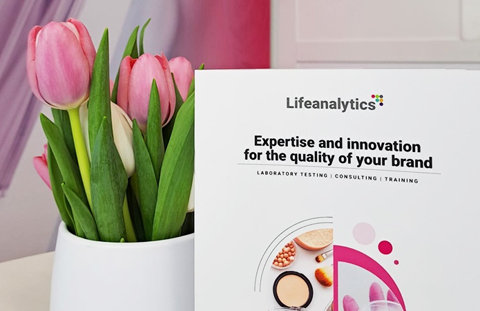 Un vaso bianco con tulipani rosa su un tavolo, fotografato accanto a un opuscolo Lifeanalytics con un'immagine di copertina di prodotti dedicati al make-up e analisi di laboratorio