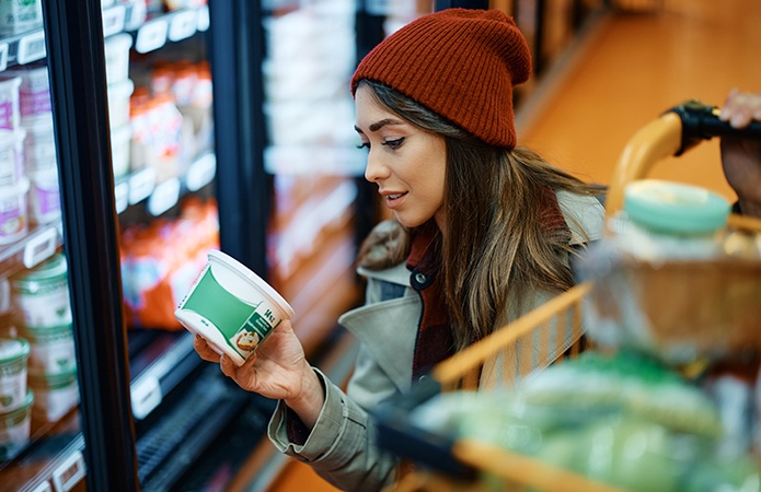 Donna giovane al supermercato mentre esamina una confezione di alimento refrigerato davanti a un banco frigo, con carrello della spesa accanto, simbolo dell'articolo del blog della business unit Food dedicato all'attenzione alle etichette con dicitura “artigianale” negli alimenti e cosa cambia con la nuova normativa