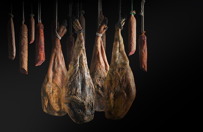 Prosciutti crudi stagionati e diversi salumi sospesi tramite corde durante il processo di stagionatura, illuminati su sfondo nero, simbolo dell'articolo del blog della business unit Food di Lifeanalytics dedicato ai nitriti e nitrati negli alimenti: cosa sono, rischi, limiti e analisi